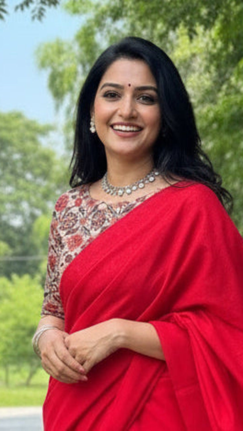 A woman standing on a pathway, wearing a vibrant solid red cotton saree with a printed blouse piece and silver jewelry.
